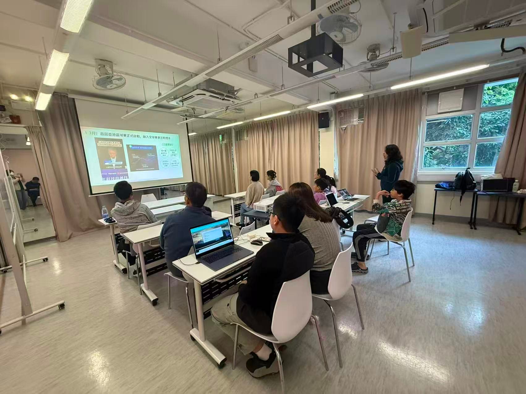 An AI learning classroom viewed from the back, with participants seated at tables with laptops, facing a projection screen.